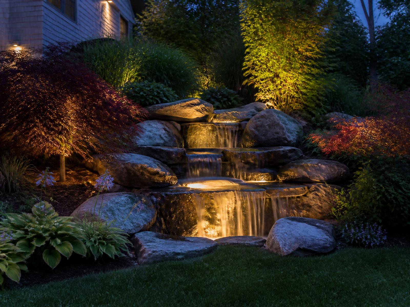 Illuminated pondless waterfall with stacked boulders and Japanese maples